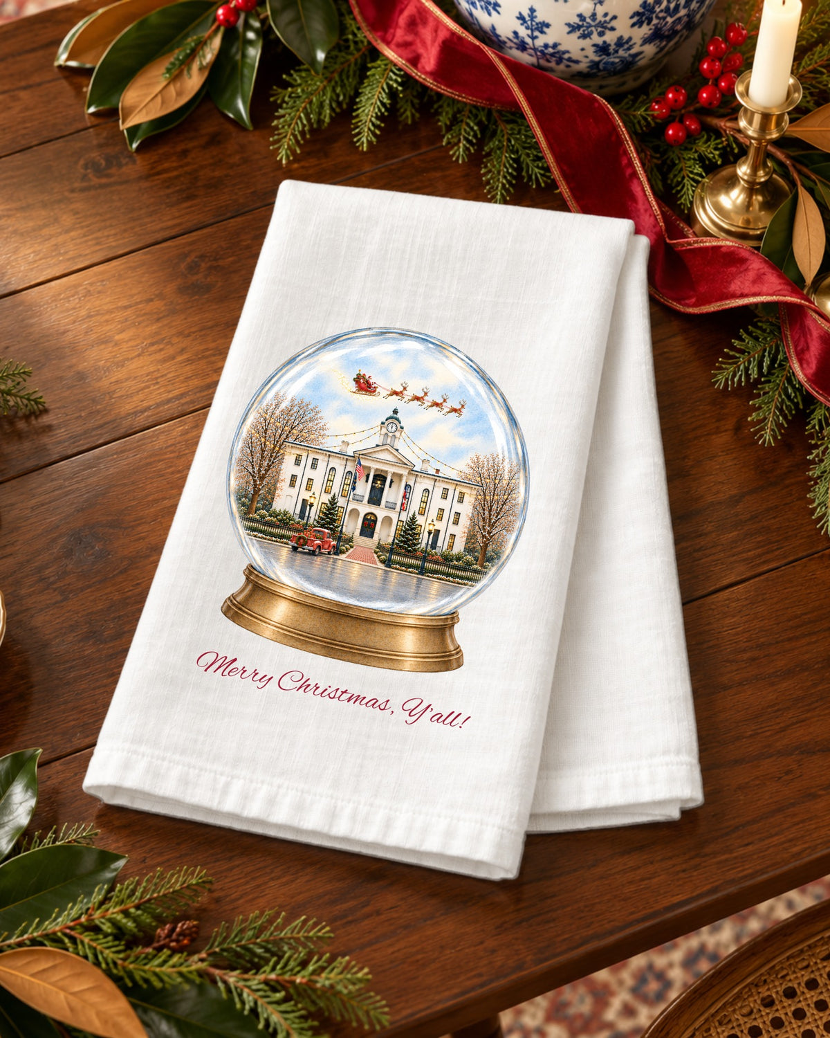 Merry Christmas, Y'all! snow globe tea towel styled in a holiday scene, showing the Oxford courthouse inside a snow globe with Santa's sleigh and a red truck.