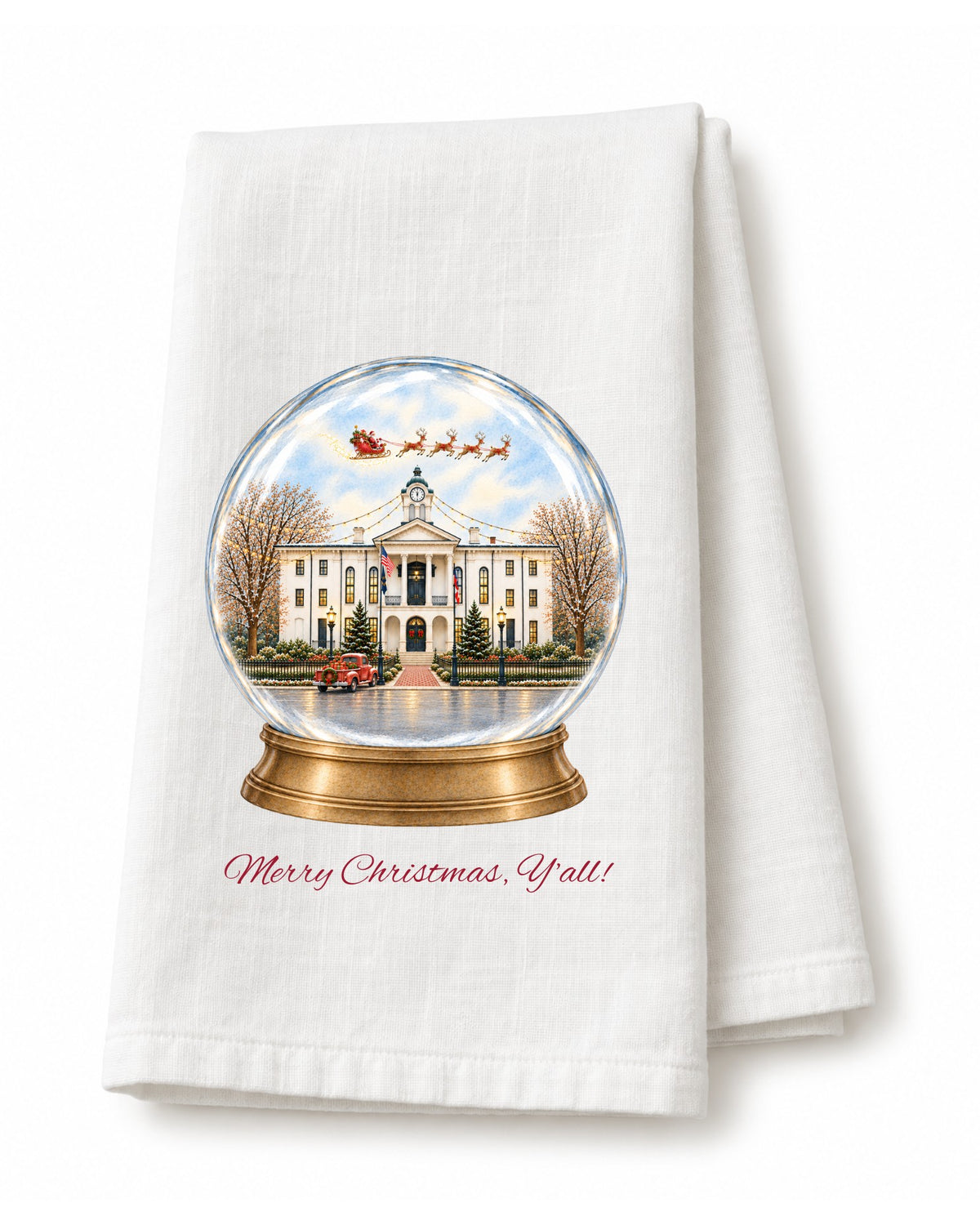 White flour sack tea towel folded, printed with a snow globe of the courthouse on the Square in Oxford with Santa's sleigh overhead, a red truck out front, and Merry Christmas, Y'all! in red script.